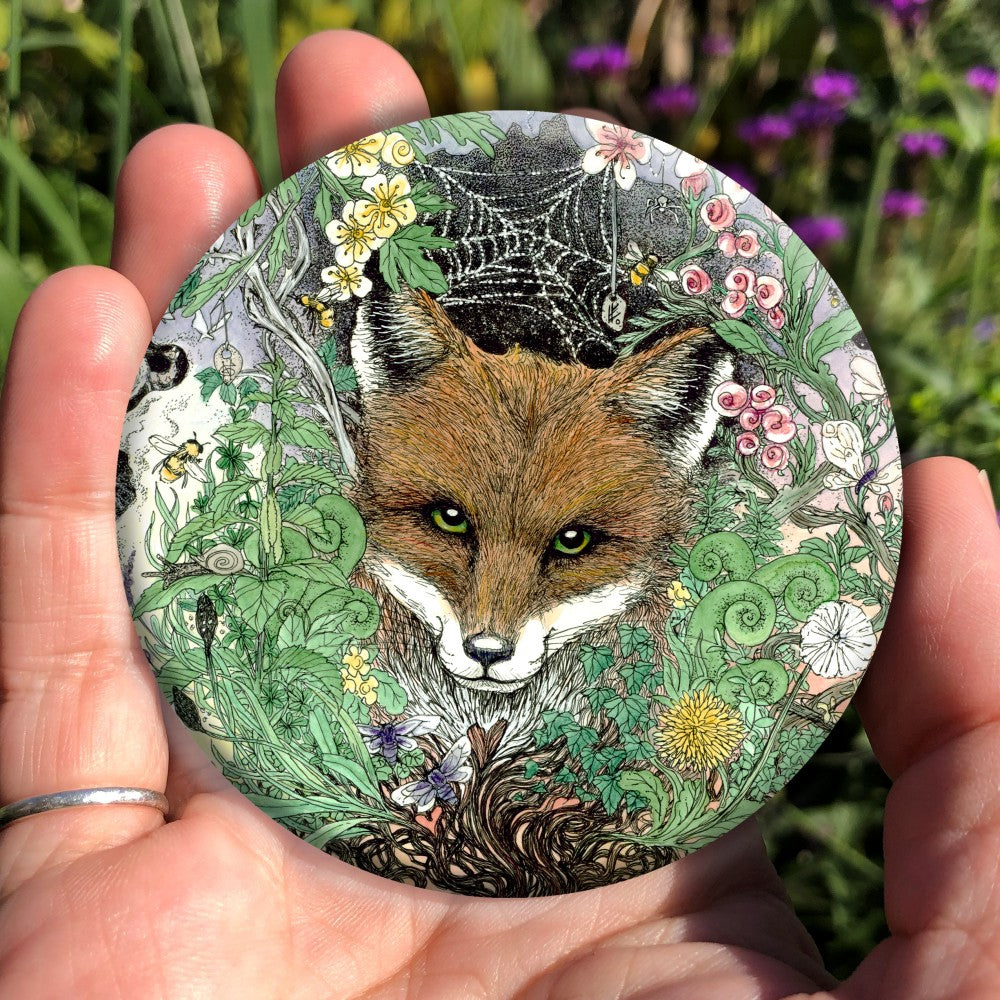 The head of a red fox looks towards the viewer. He's surrounded by a mass of leaves and flowers which rise out from a twisted tree trunk and tangled branches.