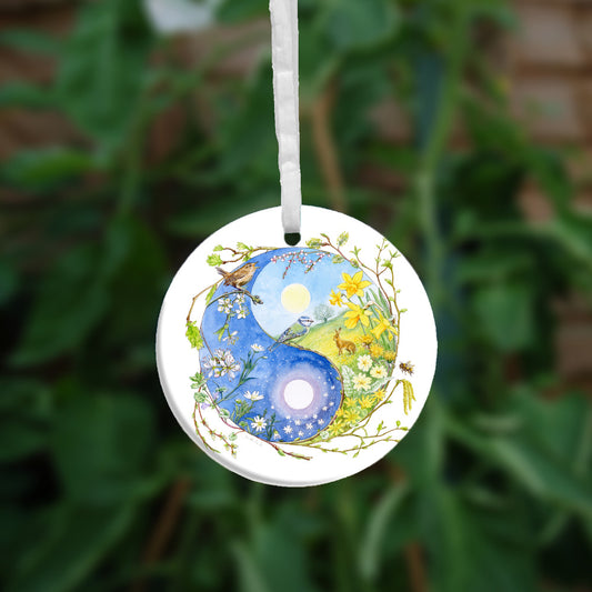 A Yin-Yang shape is surrounded by sprouting branches of native trees. The left hand half of the shape is filled with blue sky, and a large, watery sun forms the traditional 'dot'. White blossom and spring flowers overlay the sky, and a wren sings from a branch at the top. The right hand half features a hare sitting in a grassy field in a sunny day. A pale yellow sun forms the 'dot'. Yellow flowers overlay the scene. A blue tit sits on a branch forming the curve between the two sections.