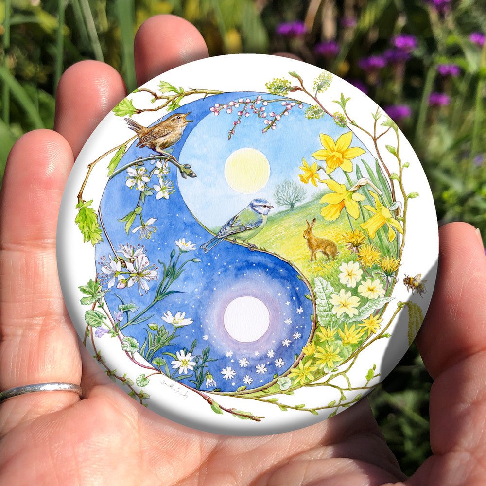 A Yin-Yang shape is surrounded by sprouting branches of native trees. The left hand half of the shape is filled with blue sky, and a large, watery sun forms the traditional 'dot'. White blossom and spring flowers overlay the sky, and a wren sings from a branch at the top. The right hand half features a hare sitting in a grassy field in a sunny day. A pale yellow sun forms the 'dot'. Yellow flowers overlay the scene. A blue tit sits on a branch forming the curve between the two sections.