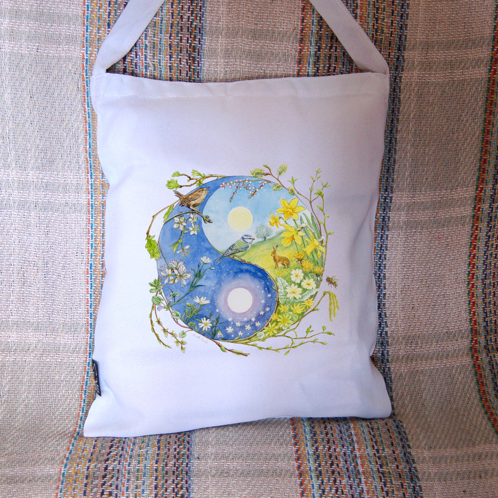 A Yin-Yang shape is surrounded by sprouting branches of native trees. The left hand half of the shape is filled with blue sky, and a large, watery sun forms the traditional 'dot'. White blossom and spring flowers overlay the sky, and a wren sings from a branch at the top. The right hand half features a hare sitting in a grassy field in a sunny day. A pale yellow sun forms the 'dot'. Yellow flowers overlay the scene. A blue tit sits on a branch forming the curve between the two sections.