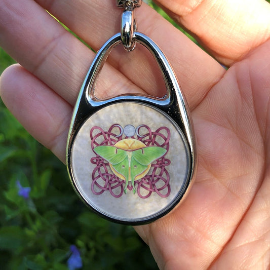 A deep pink Celtic-style knotwork fills the design. On top of the knotwork, in the centre, a Luna Moth sits on a yellow disc. There's a silver triple moon above.