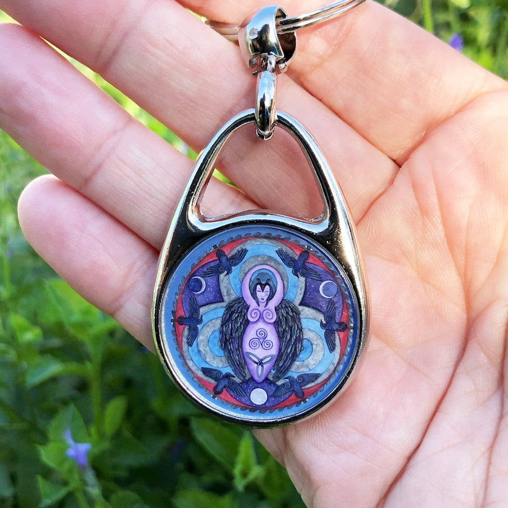 A raven stands behind a purple Goddess figure, its huge wings folded down her sides. She stands in the centre of a ring. Ravens fly around the inside of the ring, with each aspect of the triple moon also present. Colours are purple, red, and blue.